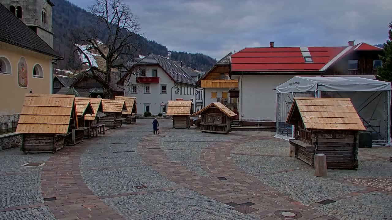 A person walks across a cobblestone town square surrounded by small wooden huts and buildings, under an overcast sky in a mountainous region.