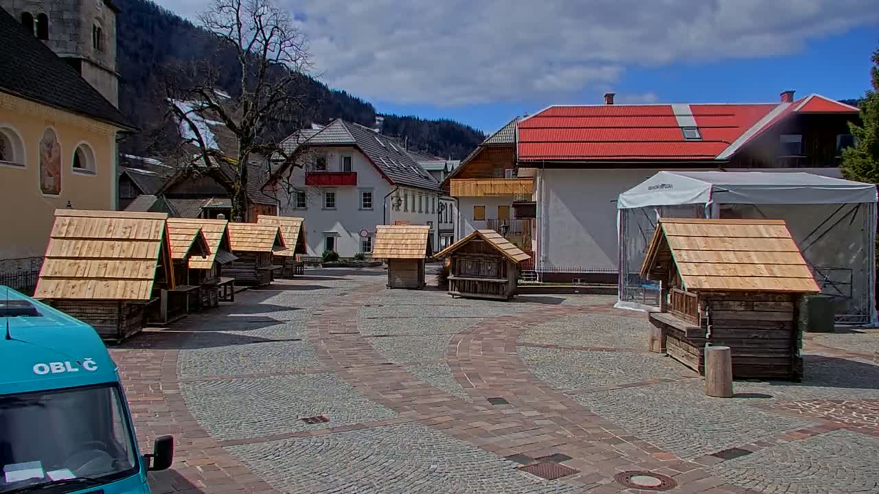 A town square with several small wooden stalls, buildings with red roofs, and mountains in the background under partly cloudy skies.