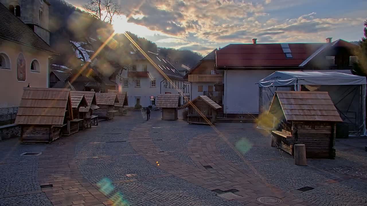 A cobblestone town square is filled with several small wooden structures under a partly cloudy sky, with the sun shining brightly behind a nearby mountain range.