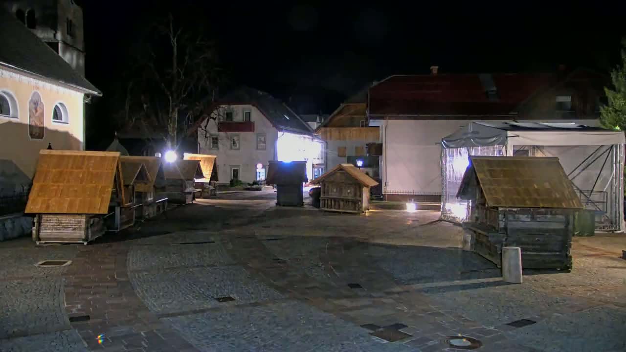 At night, several small wooden structures with gabled roofs stand in a cobblestone town square near buildings, one of which is a church.
