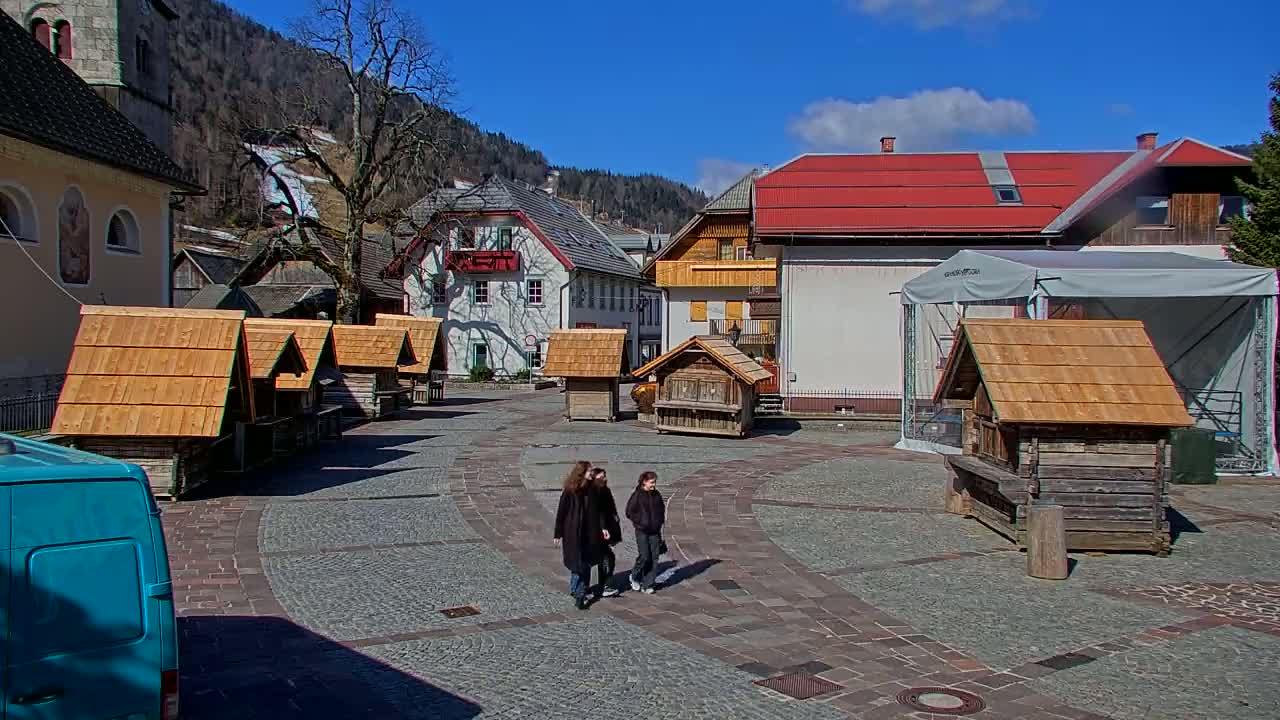 A sunny day in a European village square features several small wooden structures, two people walking, and buildings with red roofs in the background.