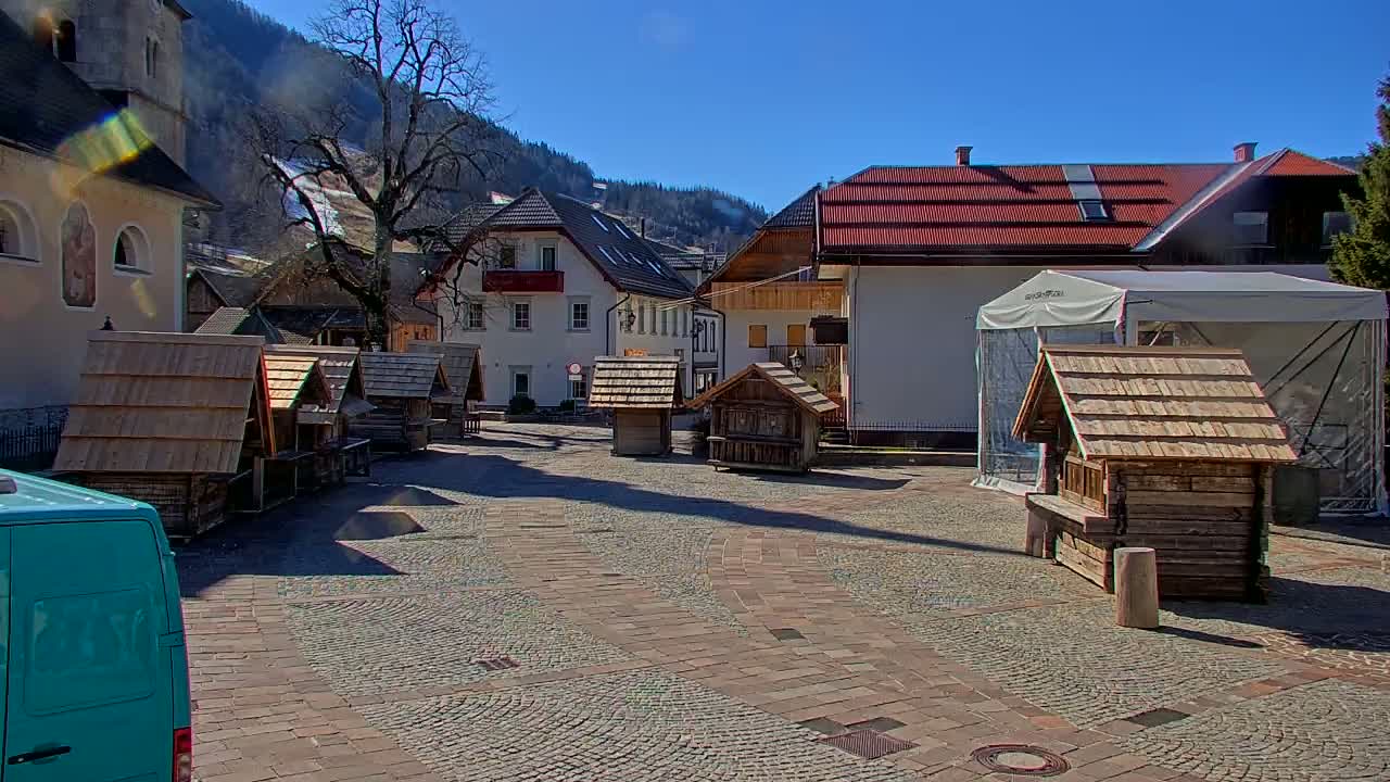 A cobblestone town square, under a clear blue sky, features several small wooden structures and buildings, with mountains in the background.