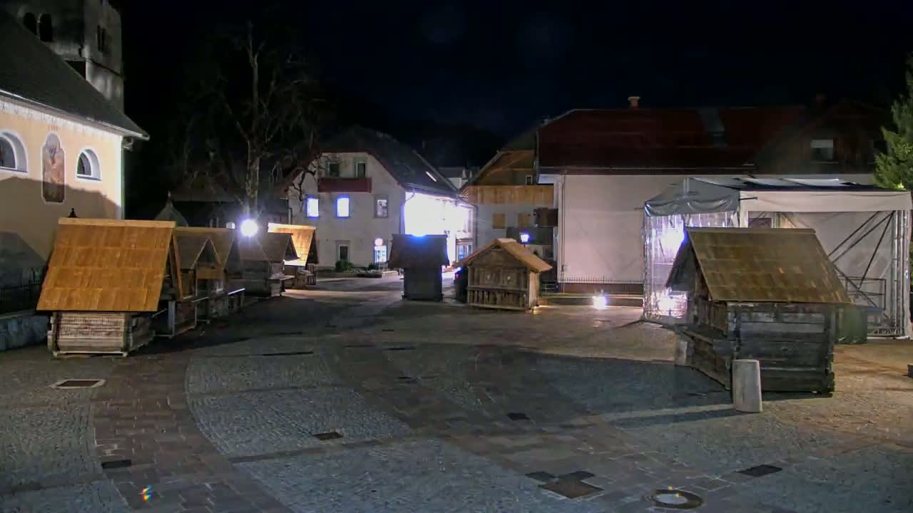 A nighttime view of a town square with several small wooden structures illuminated, near buildings with exterior lighting.