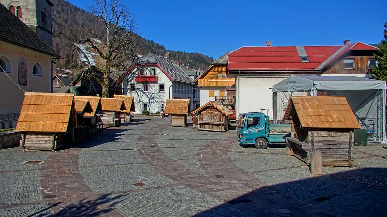 A cobblestone town square, under sunny skies, features several small wooden structures, a light blue truck, and buildings with red roofs against a backdrop of snow-dusted mountains.
