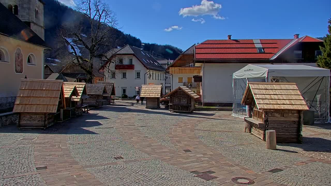 A cobblestone town square, under a mostly sunny sky, features several small wooden structures and buildings with red roofs, backed by a mountain range.