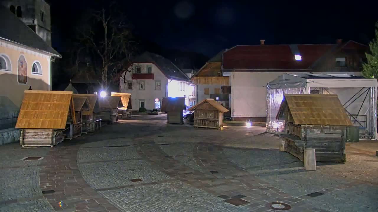 At night, several small wooden structures with thatched roofs sit in a cobblestone town square next to buildings with lights on.