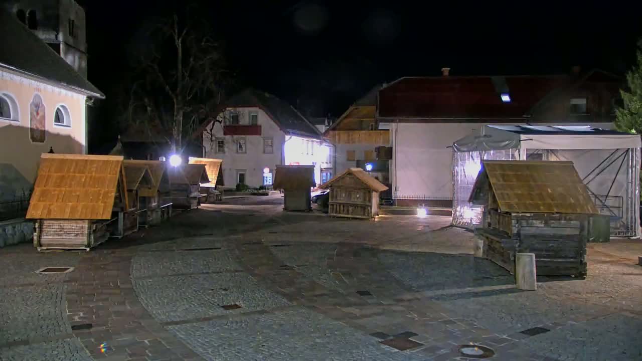 A nighttime scene shows several small wooden structures with pitched roofs in a paved square, illuminated by artificial lights, with buildings and a large tent in the background.