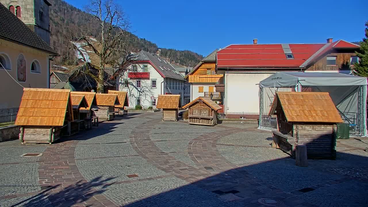 A sunny day reveals a cobblestone town square with several small wooden structures, surrounded by buildings in a mountainous region.