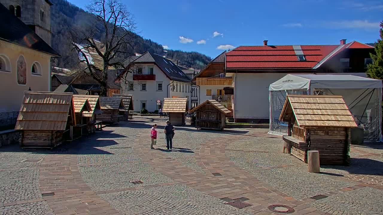 A sunny day reveals a cobblestone town square with several small wooden structures, surrounded by buildings and mountains in the background.