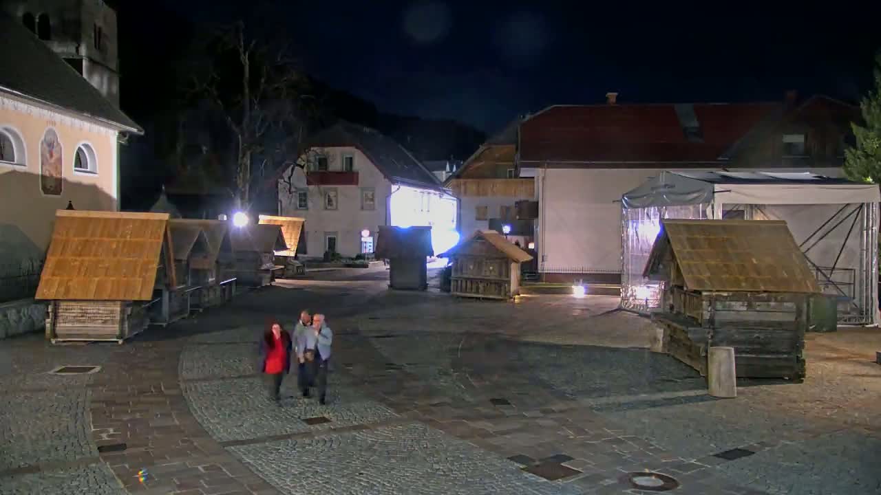 At night, three people walk across a cobblestone town square with several small wooden structures, some buildings, and a large tent.