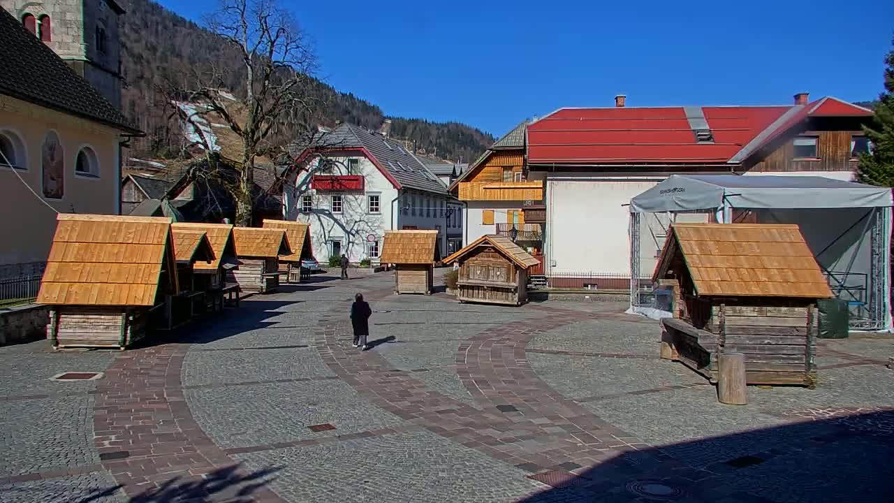 A person walks across a stone plaza in a small village nestled in a mountain valley on a sunny day,  surrounded by several small wooden structures and larger buildings.