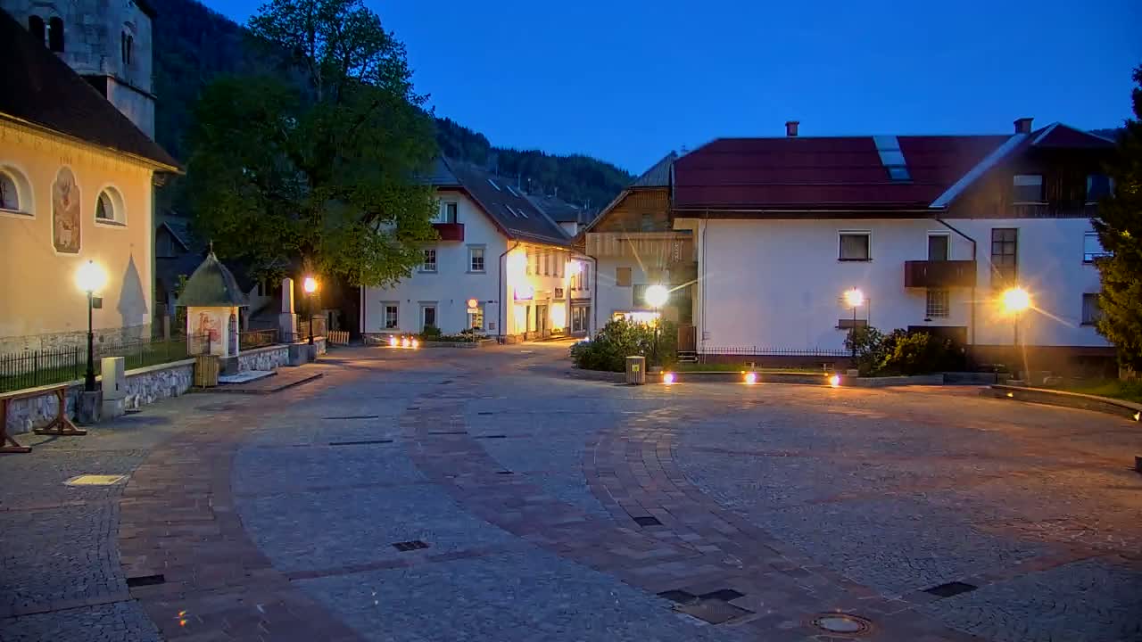 A town square at dusk, illuminated by streetlights, is surrounded by buildings nestled against a mountainside under a twilight sky.