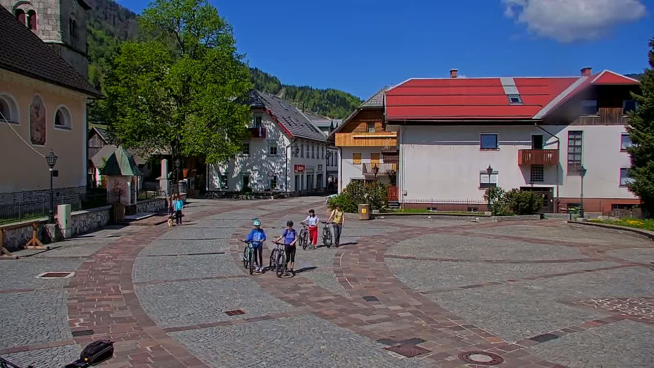 A paved town square in a mountainous region, under sunny skies, features several people on bicycles and pedestrians, with traditional European-style buildings surrounding it.