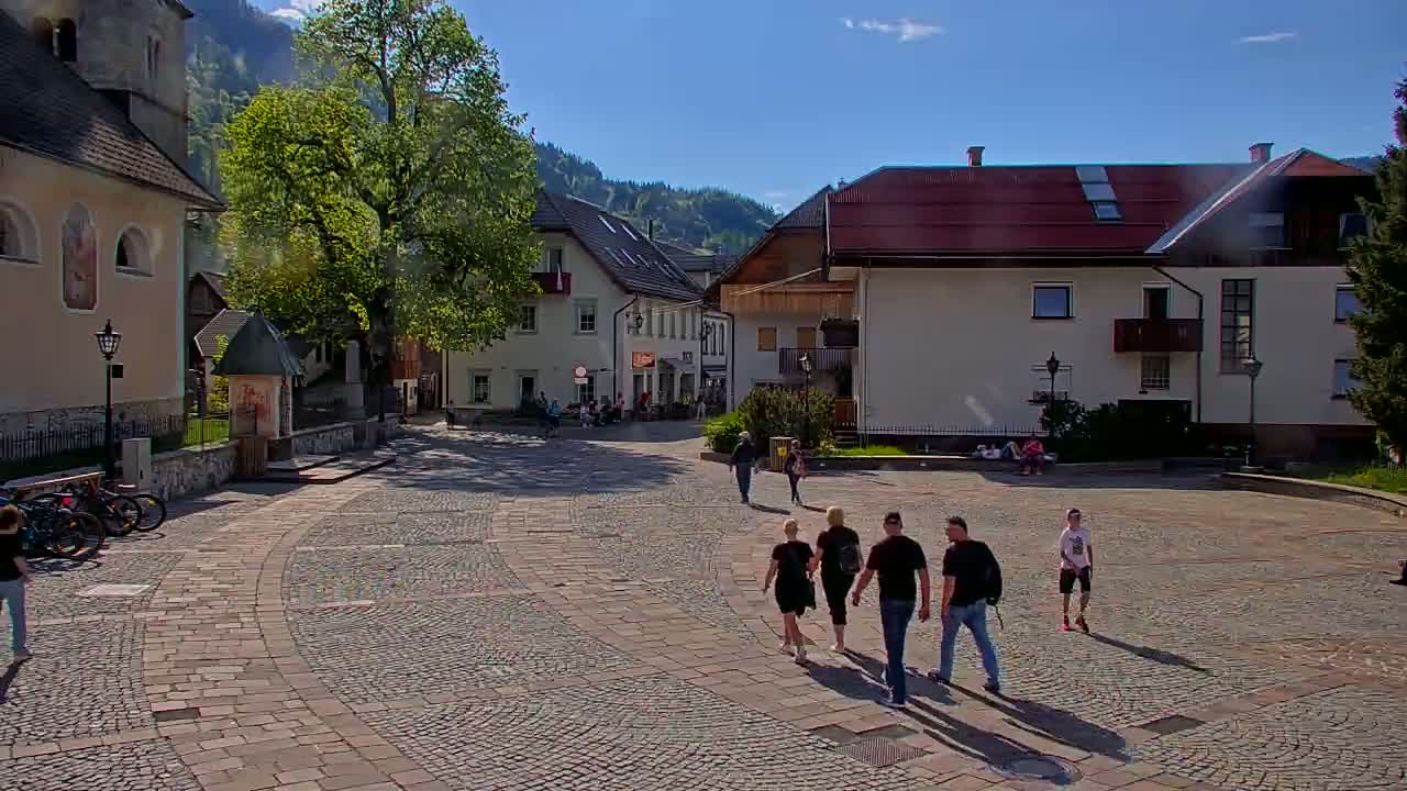 A cobblestone town square, under a sunny sky, is surrounded by buildings, with several people walking and some sitting.