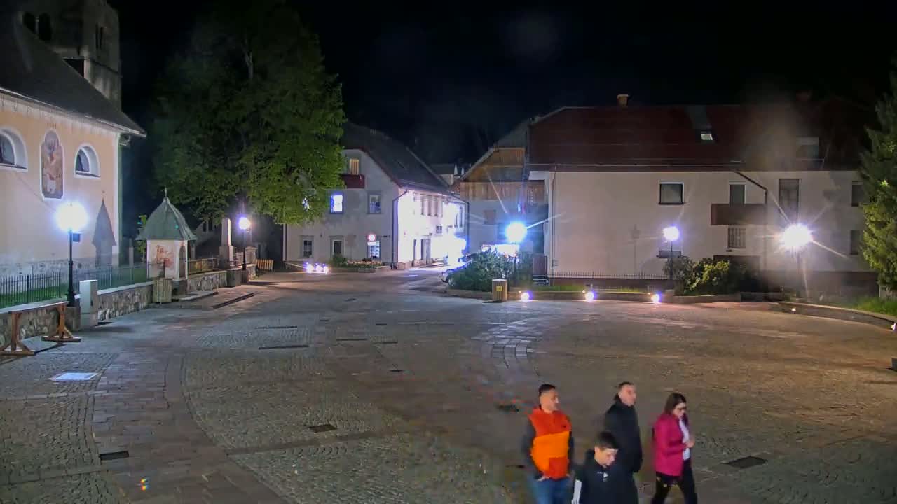 A group of people walk across a cobblestone town square at night, past buildings illuminated by streetlights.