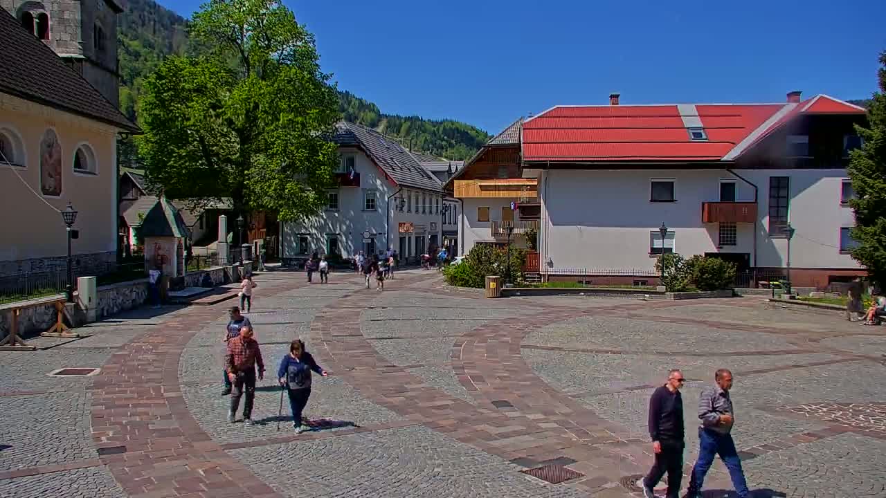 A paved town square with several people walking, surrounded by buildings including a church, under a clear, sunny sky.