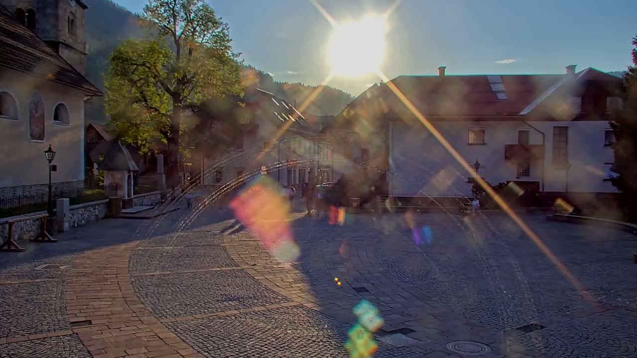 A sun-drenched cobblestone town square, surrounded by buildings and trees, with a bright sun causing lens flares.