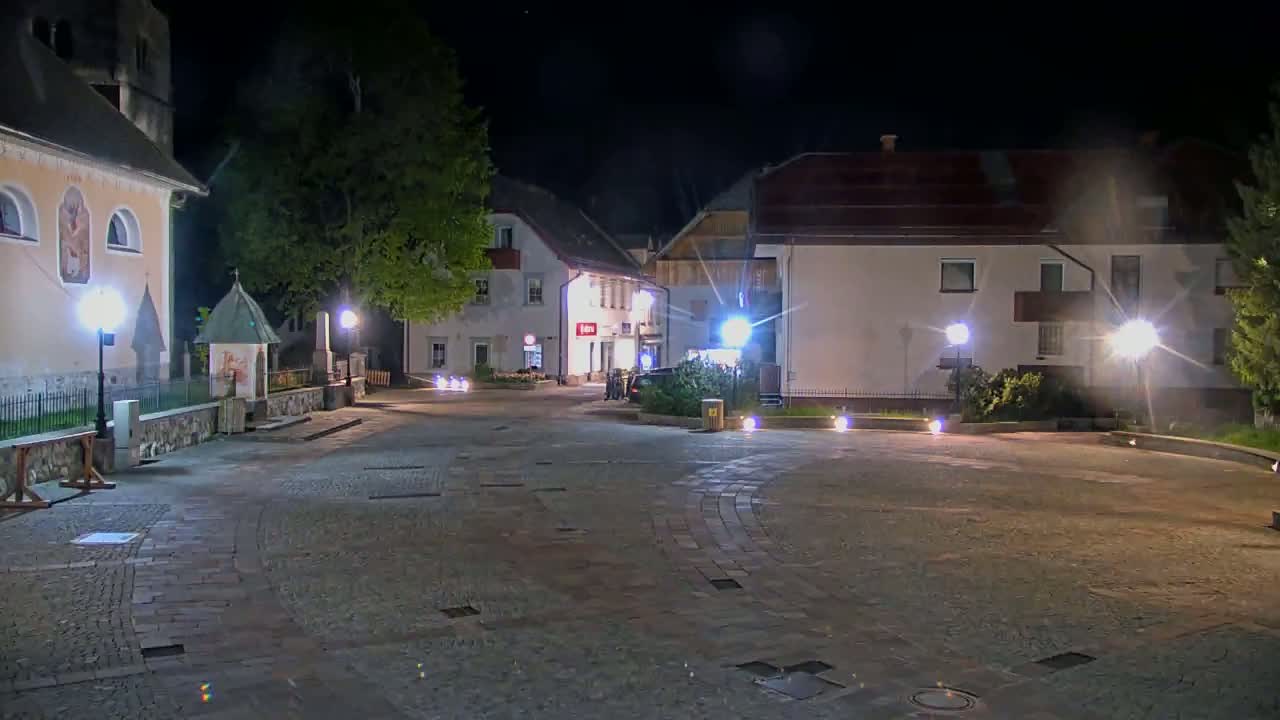 A cobblestone town square at night, illuminated by lampposts, is surrounded by buildings and trees.