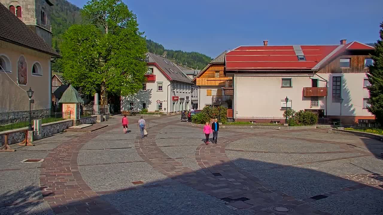 A paved town square with several buildings, including a church, and a few people walking on a sunny day.