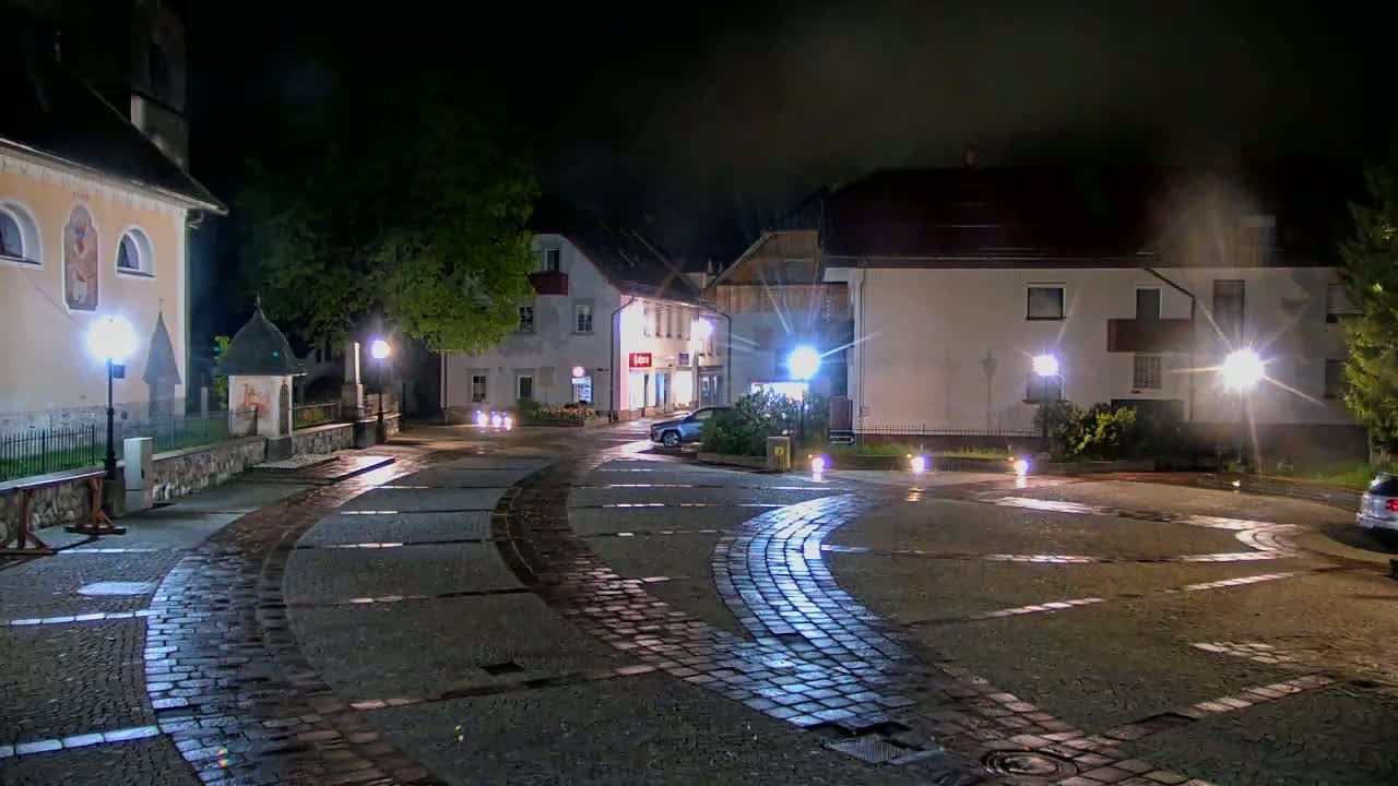 A wet cobblestone town square at night is lit by several street lamps, with buildings and a parked car visible in the background.