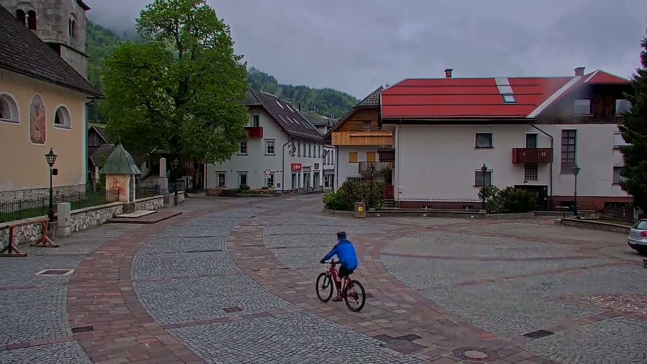 A person on a bicycle rides through a cobblestone town square, surrounded by European-style buildings under an overcast sky.