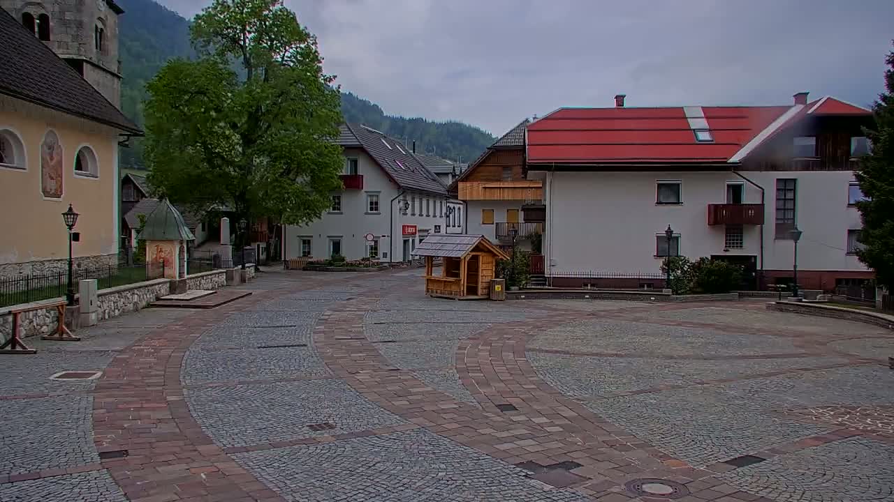 A stone plaza in a small village nestled in a valley, under an overcast sky, is surrounded by buildings, one with a red roof, and a large tree.