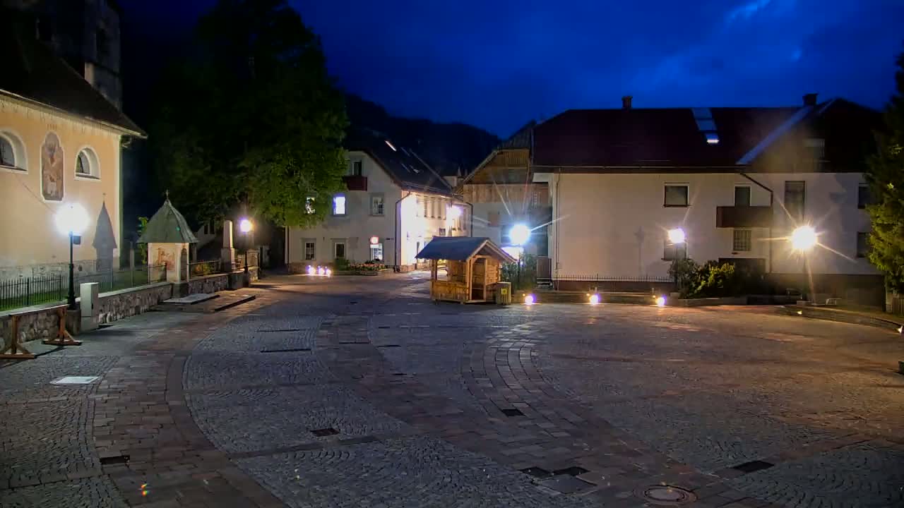 A cobblestone town square at night is illuminated by lampposts, with buildings, including a church and smaller structures, surrounding it under a dark blue twilight sky.