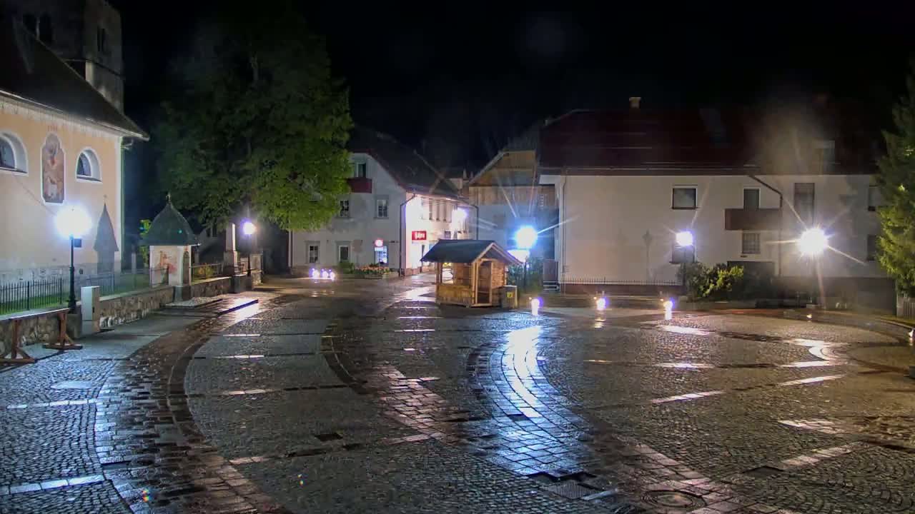 A cobblestone town square at night, illuminated by streetlights, is wet from recent rain, with buildings lining the perimeter.