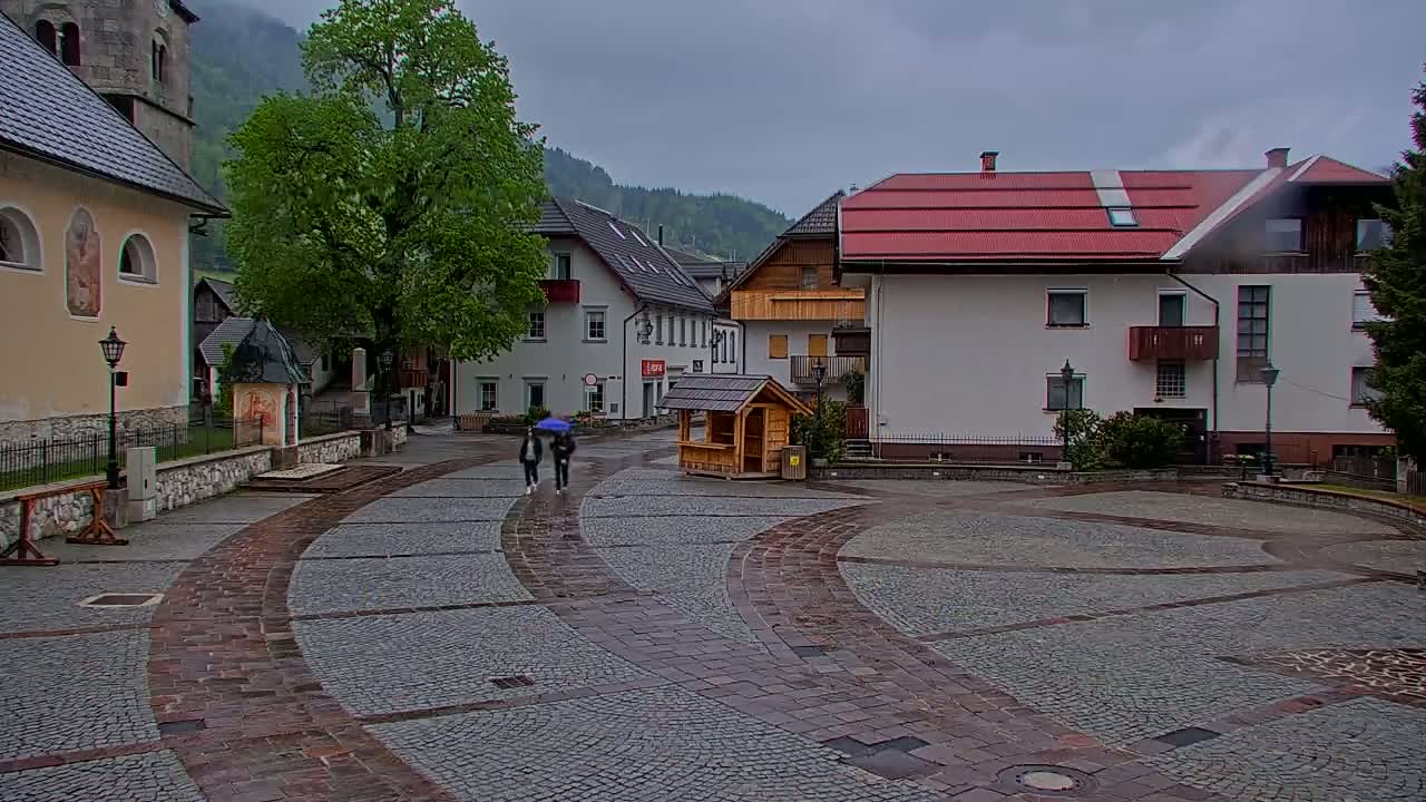 A cobblestone town square, with two people under an umbrella walking toward a small wooden structure, is surrounded by buildings in a mountainous area on a cloudy, rainy day.