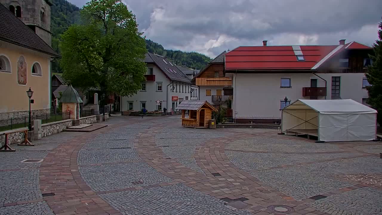 A cobblestone town square, under a cloudy sky, features buildings, a tree, a small wooden structure, and a white tent on a stage.
