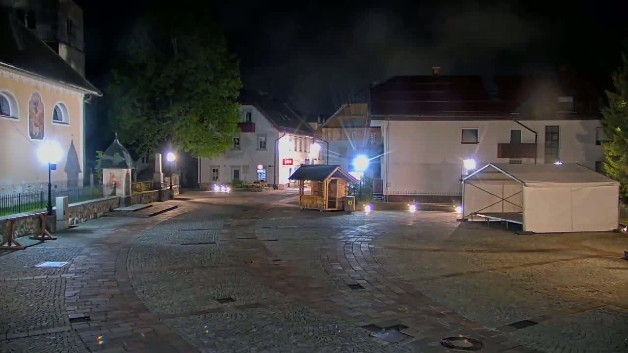 A cobblestone town square at night is lit by streetlights, featuring buildings, a small wooden booth, and a large beige tent.
