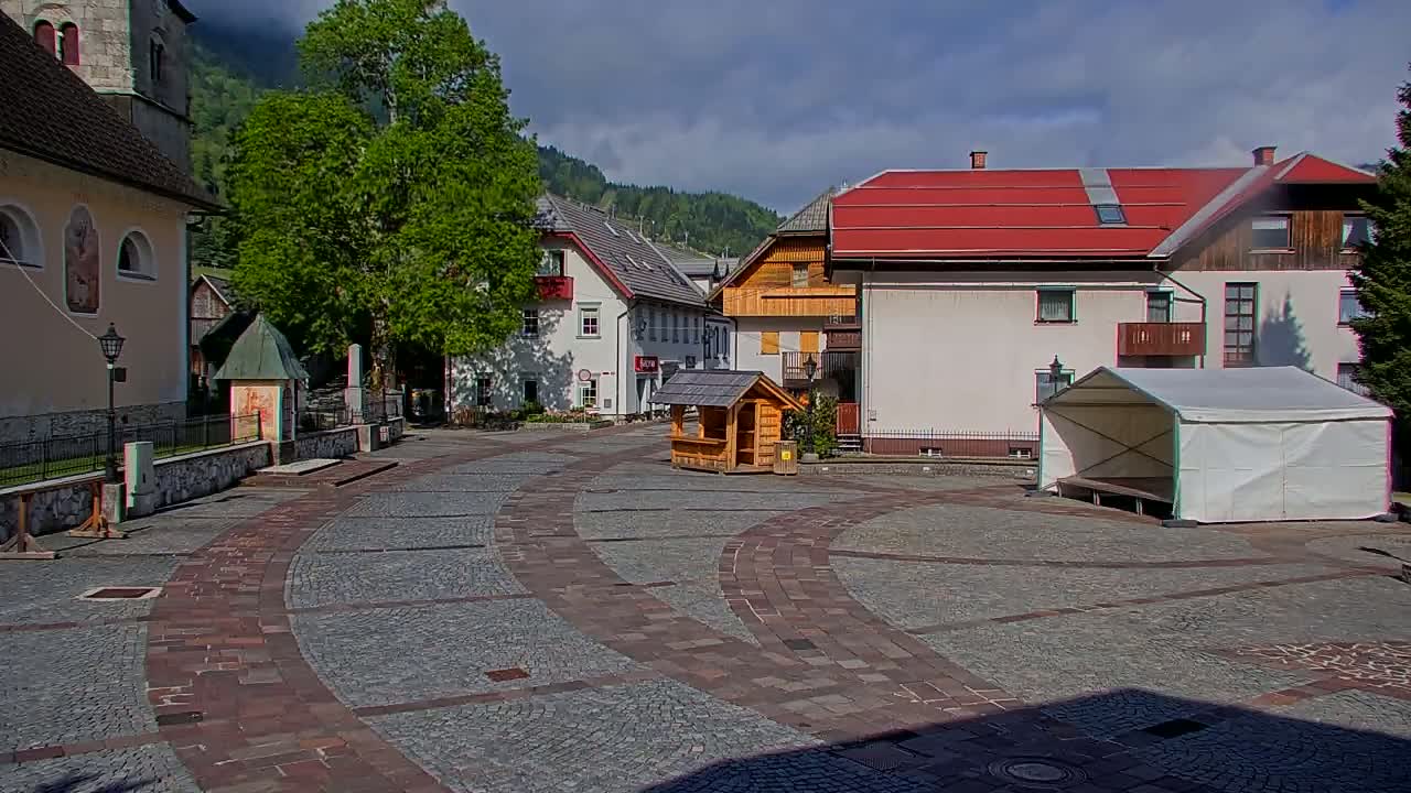 A cobblestone town square with several buildings, a small wooden structure, a white tent, and a large tree under a partly cloudy sky.