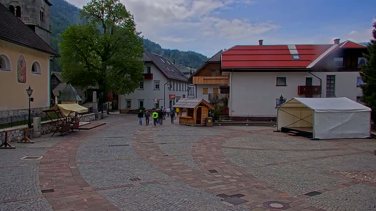 A cobblestone town square, under partly cloudy skies, features several buildings, a small group of people walking, a wooden kiosk, and a white event tent.