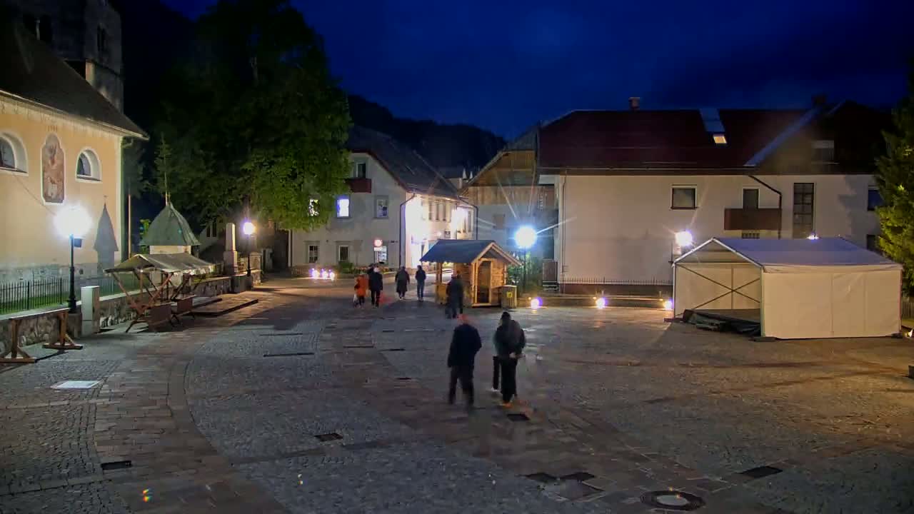 A town square at night, illuminated by lampposts, features several buildings, a small wooden structure, a white tent, and a few people walking.