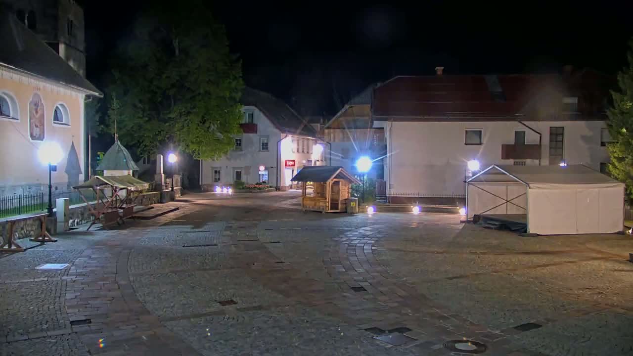 A cobblestone town square at night is illuminated by lampposts, featuring several buildings, a small wooden structure, and a white tent.