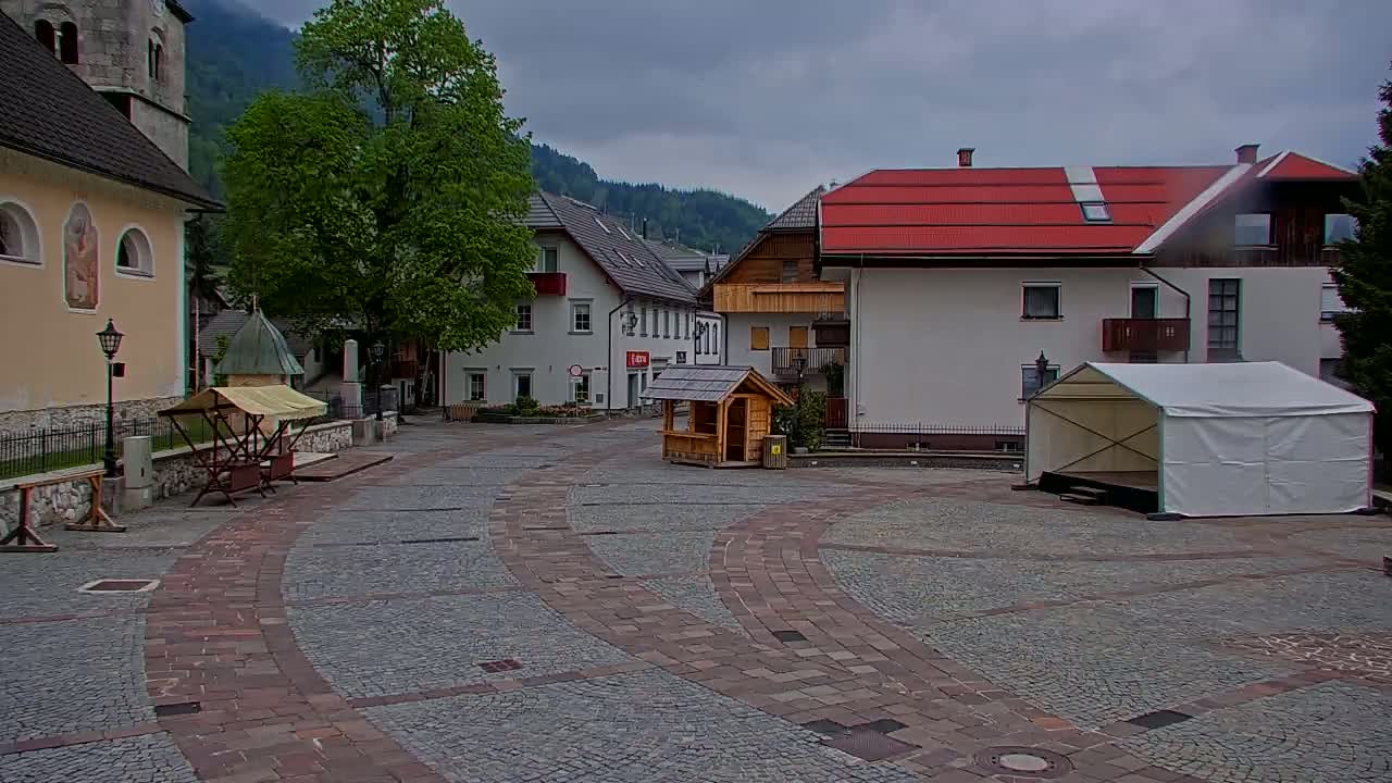 A cobblestone town square is situated between buildings, one of which is a church, under an overcast sky, with a small wooden structure and a white canopy stage in the center.