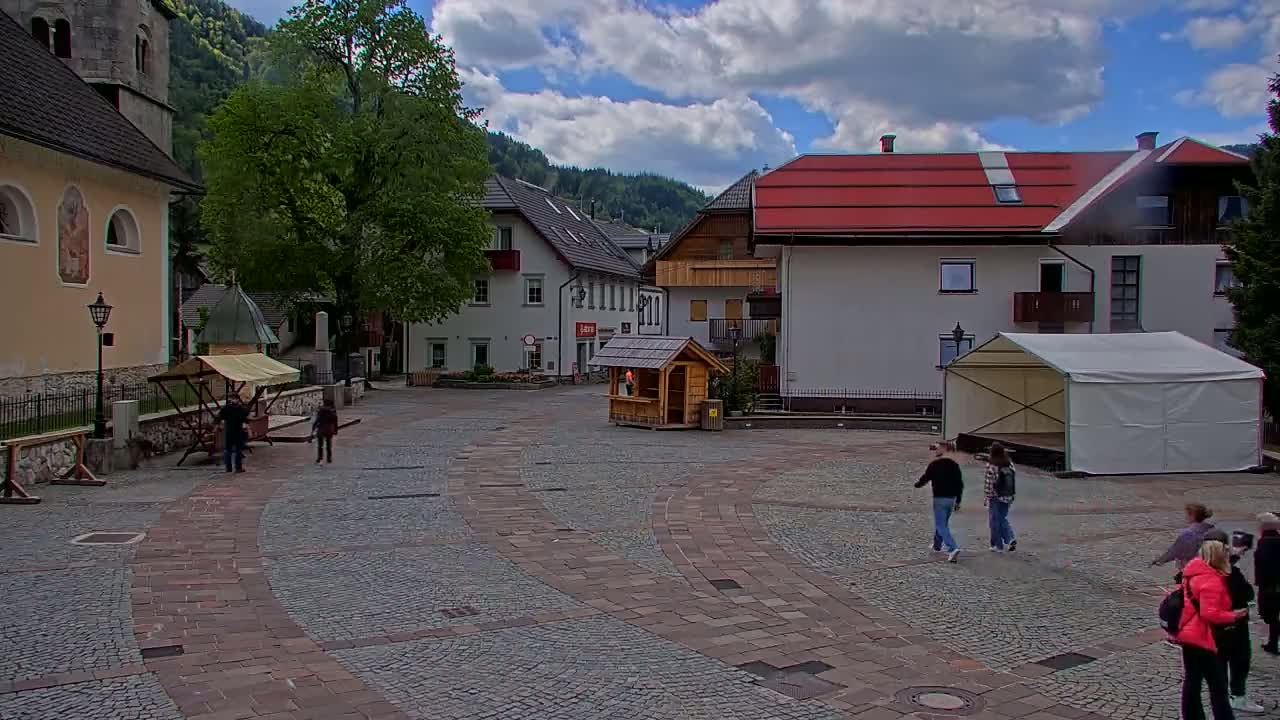 A cobblestone town square, under partly cloudy skies, features several buildings, a small wooden booth, a large white tent, and a few pedestrians.