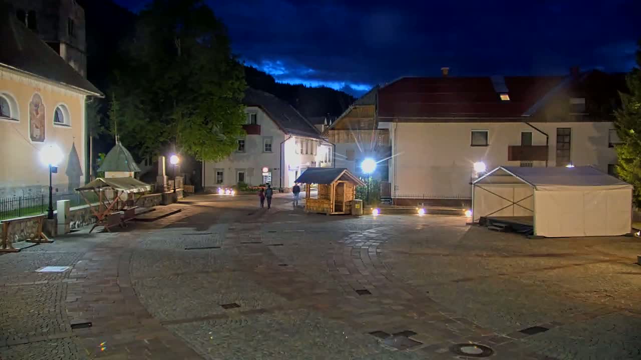 A nighttime view of a town square with several buildings, a small wooden structure, a covered stage, and a few people walking.