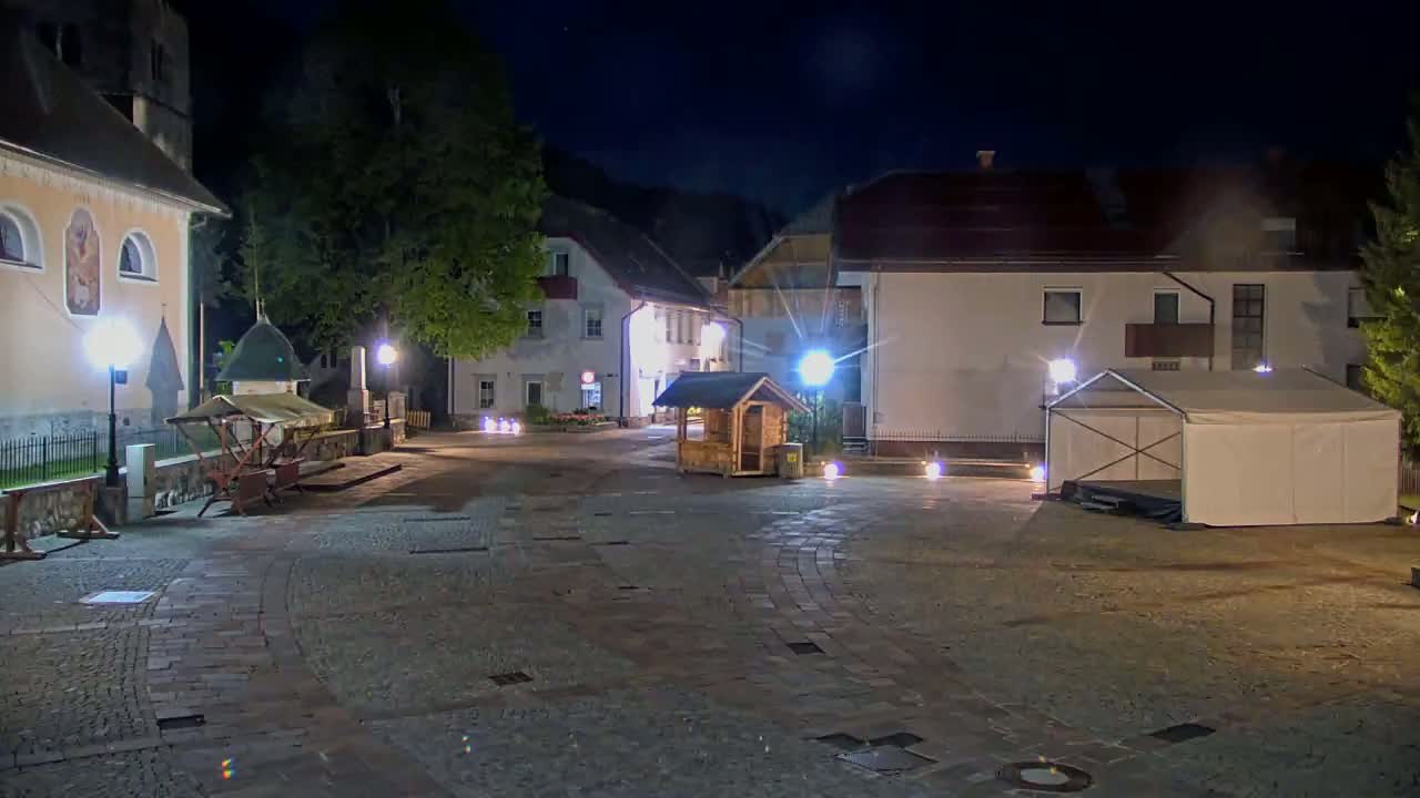 A nighttime view of a town square, illuminated by streetlights, featuring buildings, a small wooden structure, a covered stage, and a stone pavement.