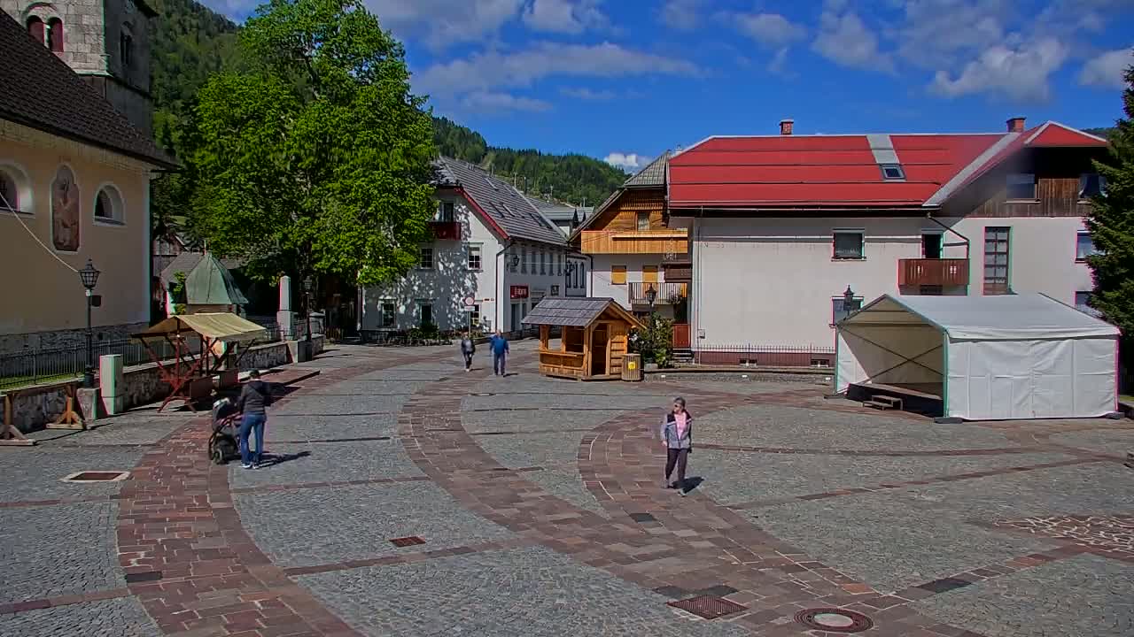A paved town square, under a sunny sky with some clouds, features several buildings, a few people walking, a small wooden booth, and a white tent.