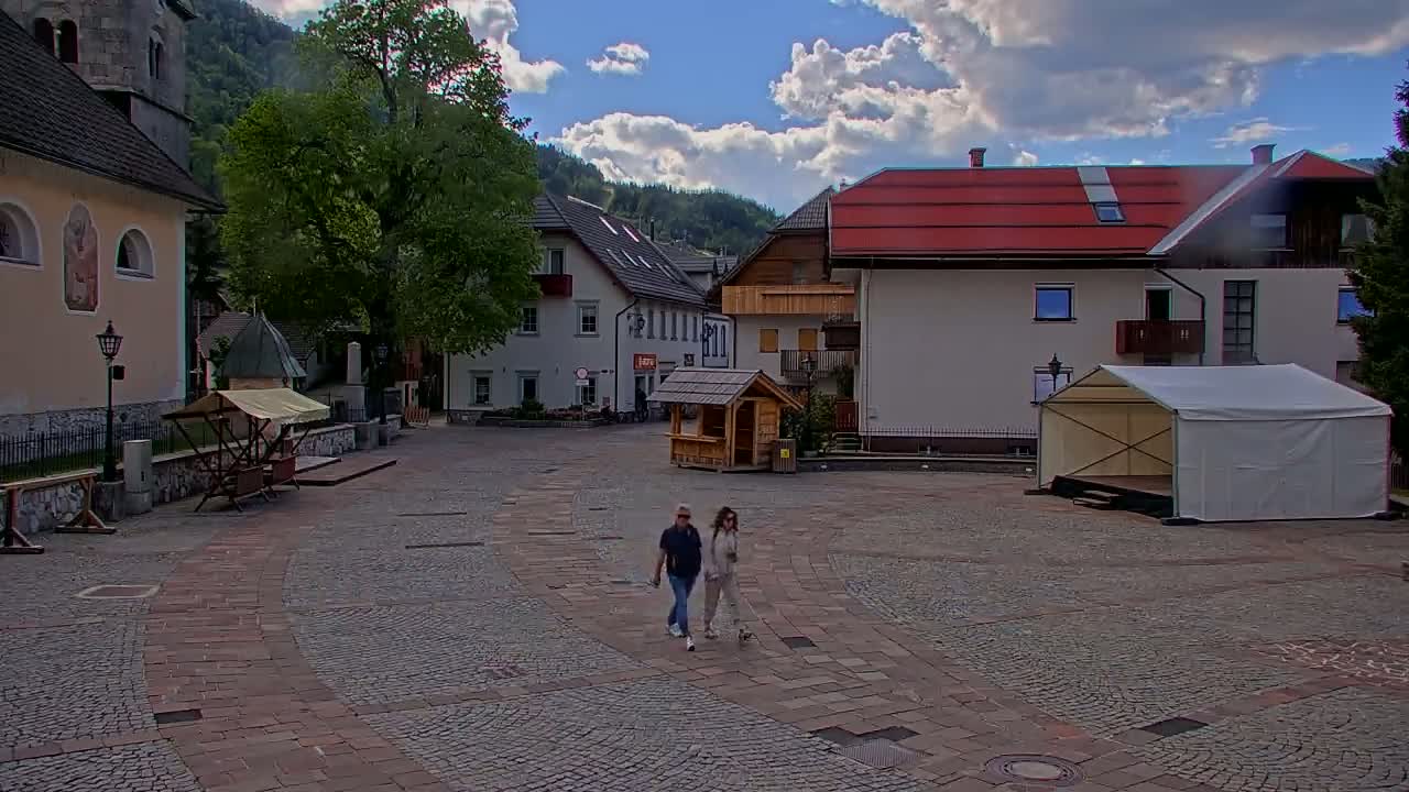 A paved town square, under partly cloudy skies, features several buildings, a small wooden kiosk, a white tent stage, and two people walking a small dog.