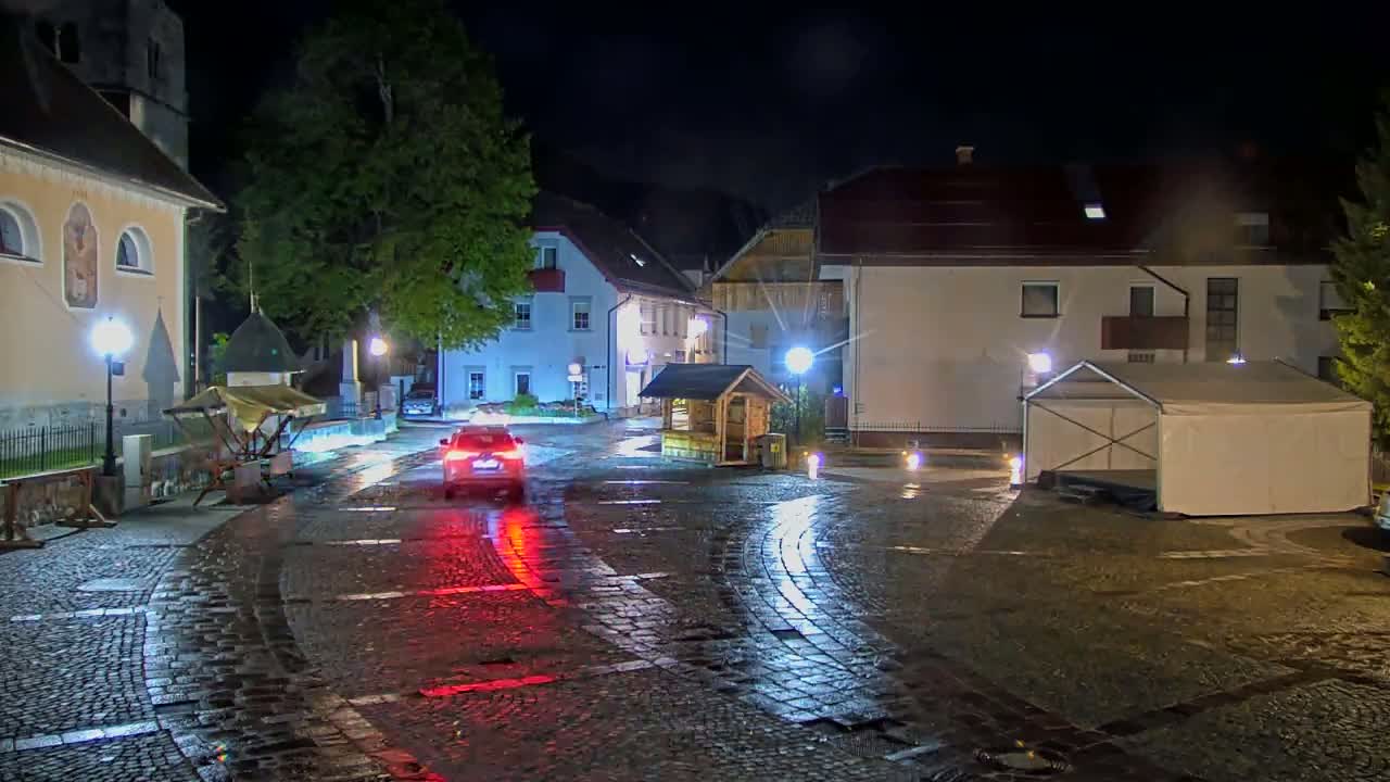 A wet cobblestone town square at night is illuminated by streetlights, with a car parked in the center and a white tent to the right.