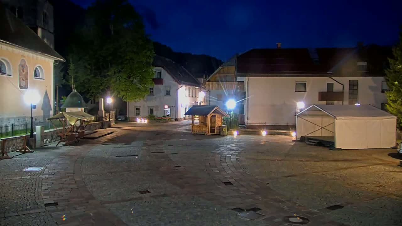 A cobblestone town square at night is illuminated by streetlights, featuring buildings, a small wooden structure, and a large white tent.
