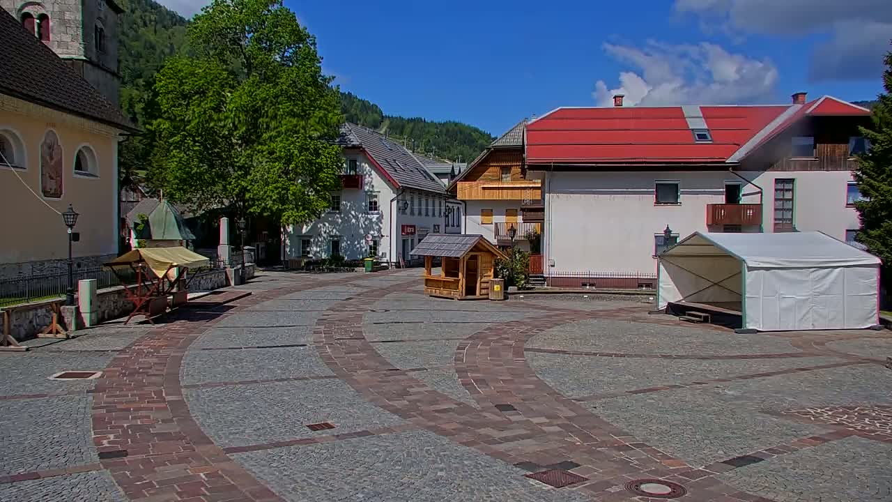 A sunny day in a cobblestone town square, surrounded by buildings including a church and a small wooden kiosk, with a white tent structure in the foreground.