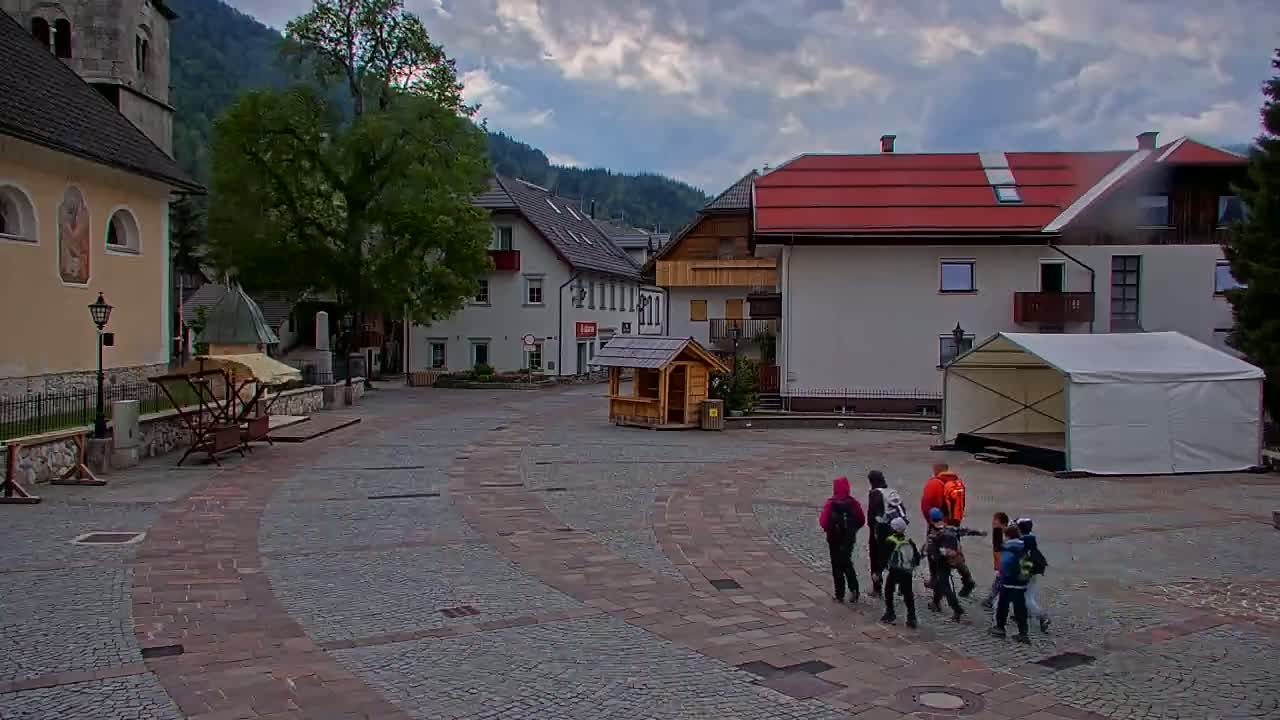 A group of people walk across a cobblestone town square under a cloudy sky, near buildings, a small wooden structure, and a white tent.
