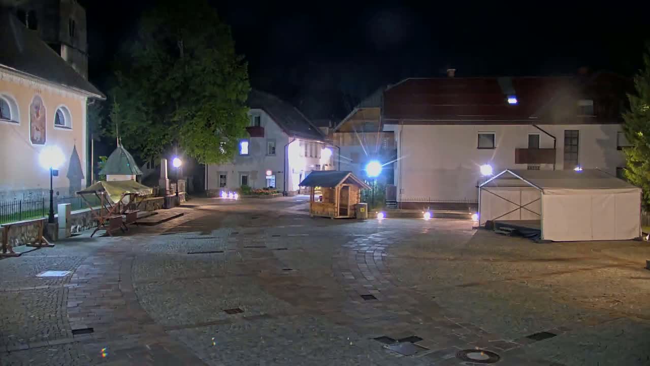 A cobblestone town square at night is illuminated by lampposts, featuring several buildings, a small wooden structure, a white tent, and a wooden stall.