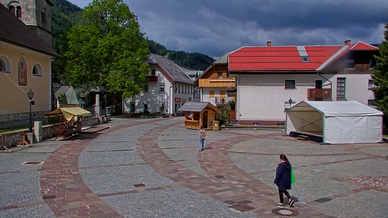 A town square with a cobblestone pavement, surrounded by buildings, including a church and a structure with a red roof, under a partly cloudy sky.