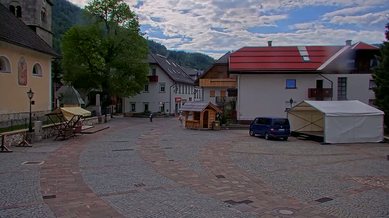 A cobblestone town square, under a partly cloudy sky, is surrounded by buildings, including a church and a small wooden kiosk, with a blue van and a white tent in the foreground.
