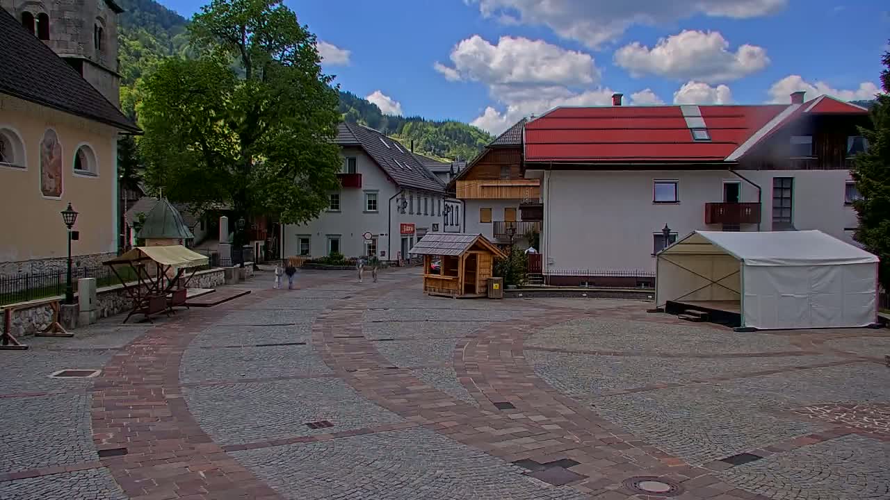 A cobblestone town square, under partly cloudy skies, features buildings of various styles, a small wooden stall, and a white tent stage.