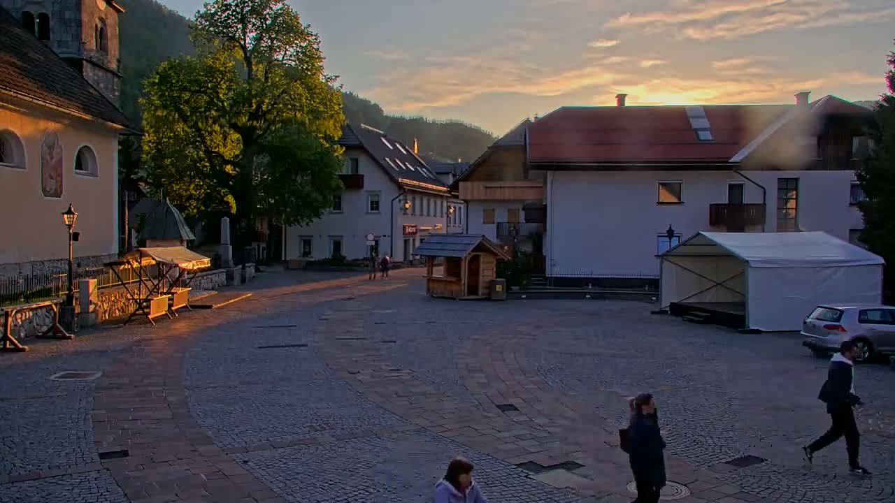 A cobblestone town square at sunset, nestled between buildings and hills, features a few pedestrians and a small, empty market stall.